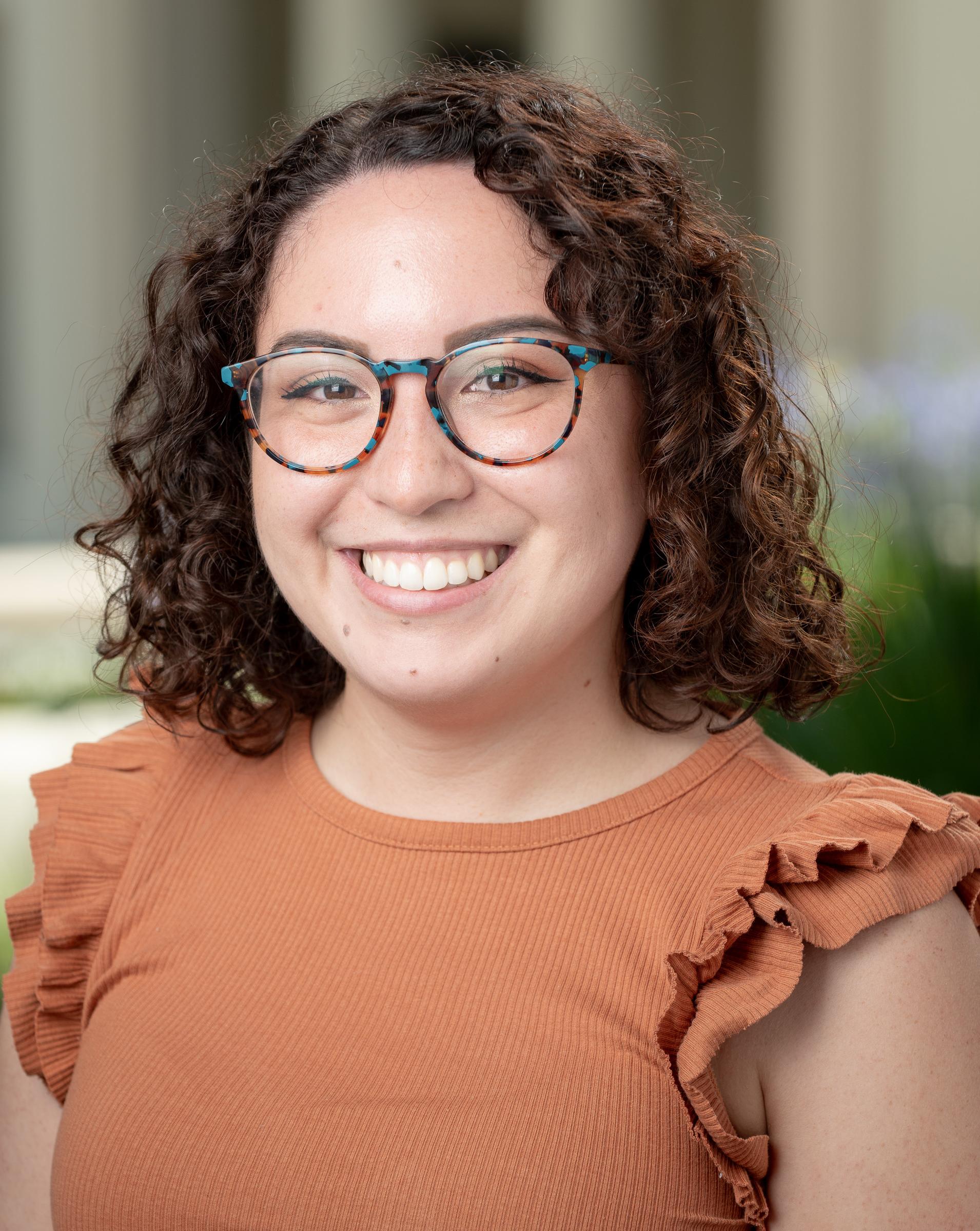 Image displays a woman with shoulder-length brunette curly hair and glasses wearing an orange shirt