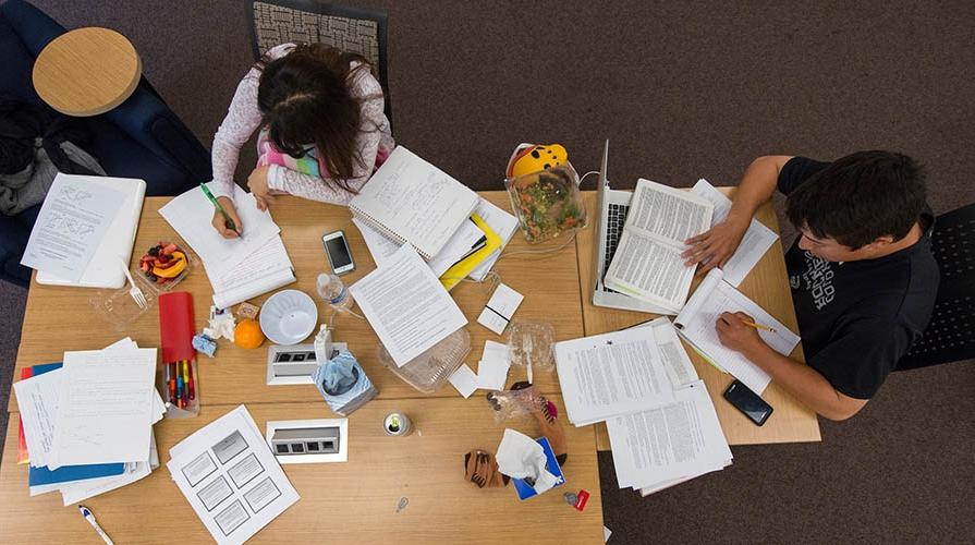 Two students study at a table surrounded by papers and notebooks