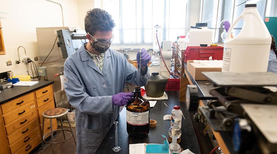 A student wearing a lab coat, gloves, and mask holds a beaker in a science lab