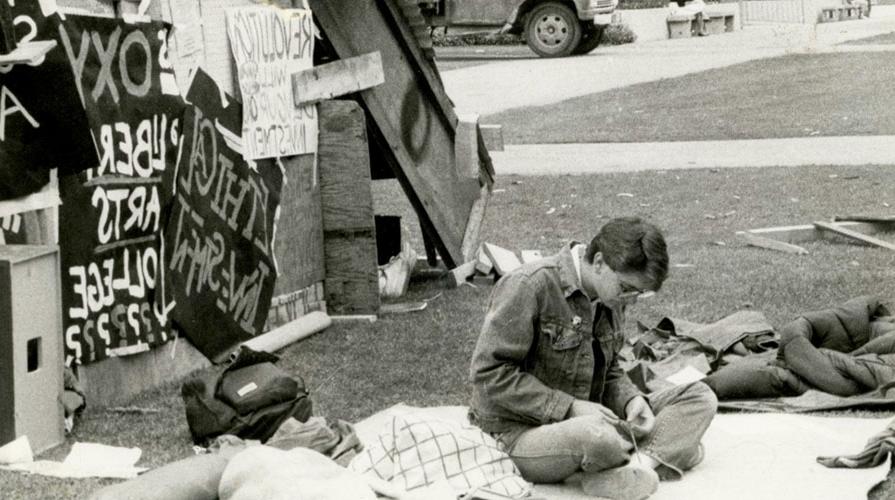 A black and white photo of an Oxy student sitting in protest with a number of signs in the background