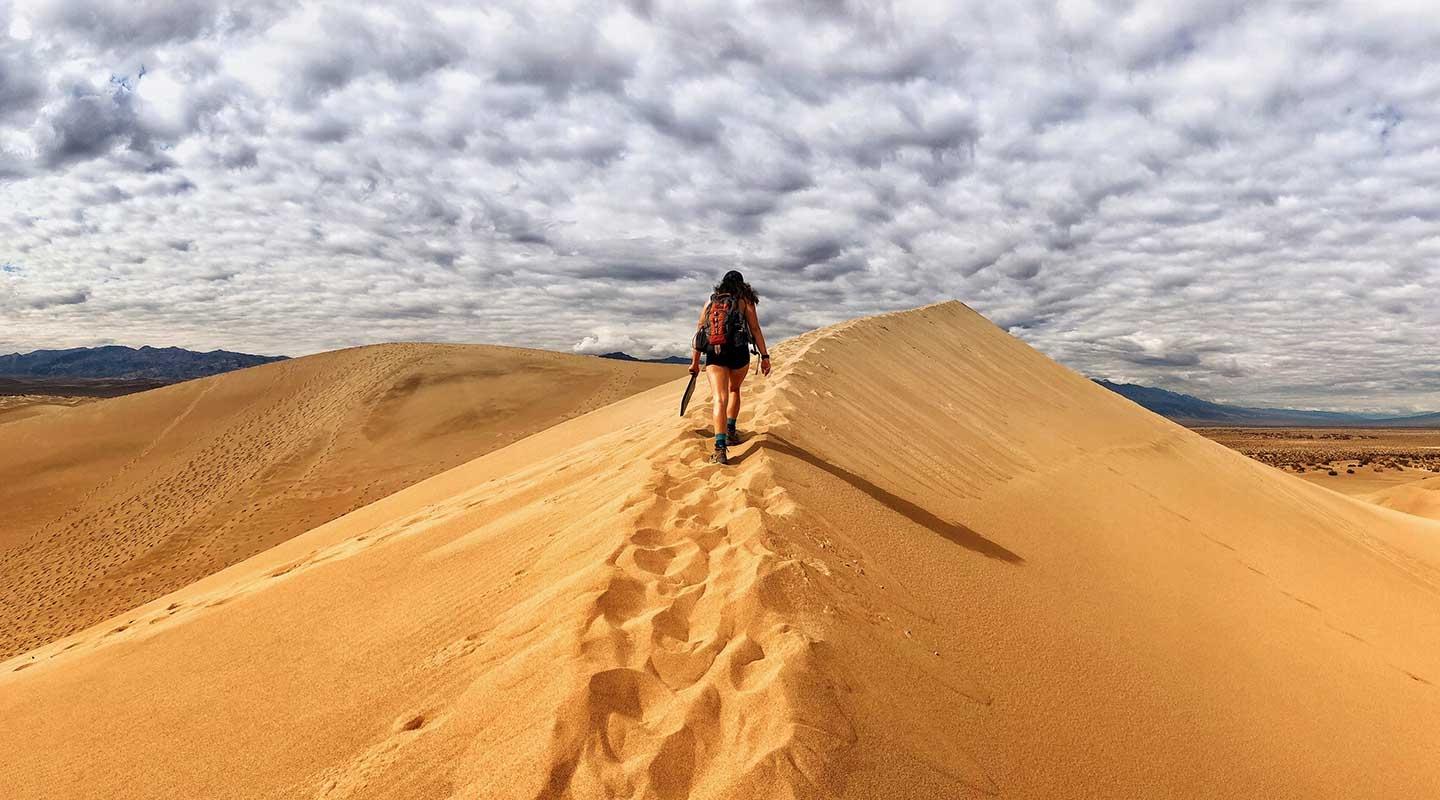 A student hiking along a sandy ridge in the desert with dramatic sky