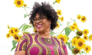 Monica White headshot in a colorful dress in front of tall sunflower plants