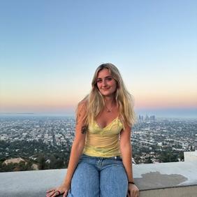 Grace is sitting on a wall with the city and sunsetting sky in background