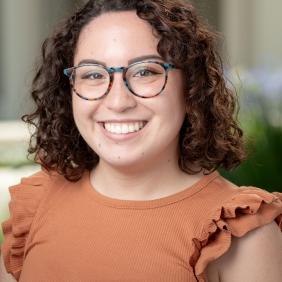 Image displays a woman with shoulder-length brunette curly hair and glasses wearing an orange shirt