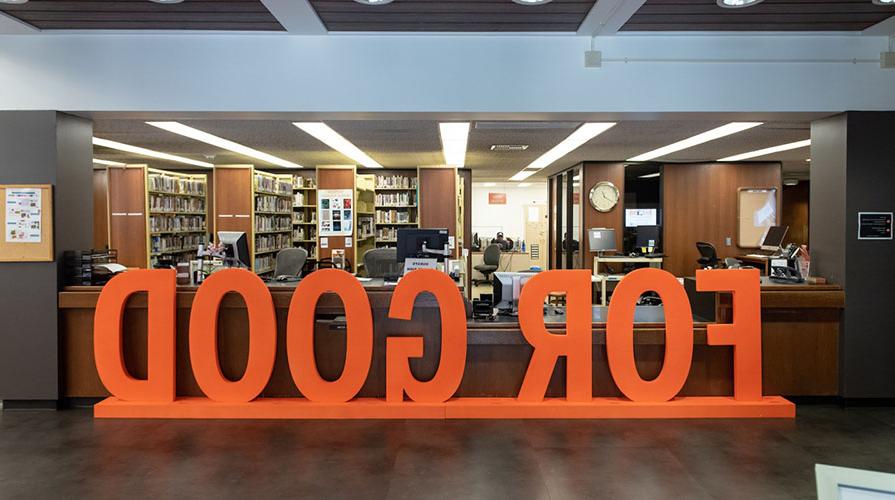 Large wooden orange sign reading "FOR GOOD" rests in front of a library circulation desk. 